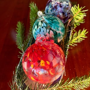May include: Three glass ornaments with a mottled, crackled finish. The ornaments are red, teal, and purple. The ornaments are sitting on a tray with a green pine branch.