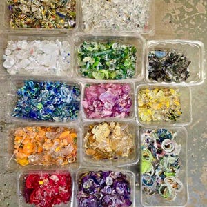 May include: An overhead shot of twelve clear plastic containers, each filled with colorful, irregularly shaped glass pieces. The colors include clear, blue, green, purple, yellow, orange, and red. The glass appears to be recycled or repurposed.