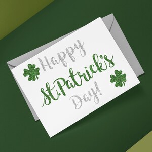 May include: A white greeting card with a gray envelope featuring the text Happy St. Patrick's Day in silver and green glitter script. Two green glitter shamrocks flank the text. The logo DnA Designed is visible in the top left corner.