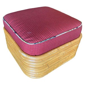May include: A square ottoman with a burgundy cushion featuring a textured pattern and white piping. The base is made of light brown, curved bamboo. This piece of furniture is suitable for a living room or bedroom.