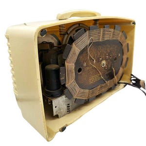 Puede incluir: Una radio antigua de color crema con su panel trasero retirado, revelando los componentes internos. La radio presenta un asa, un altavoz y una bobina grande de forma ovalada. Se ven cables y otras piezas electr&oacute;nicas en el interior.