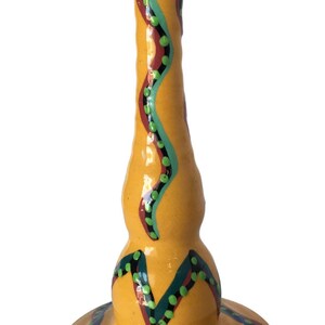 May include: A yellow ceramic candle holder with a wide base and a tall, slender neck. The holder is decorated with a colorful, abstract design featuring green, blue, red, and black lines and dots.