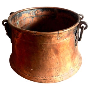 May include: A large, antique copper pot with two iron handles. The pot has a rounded bottom and a wide opening. The copper has a patina of age and wear.
