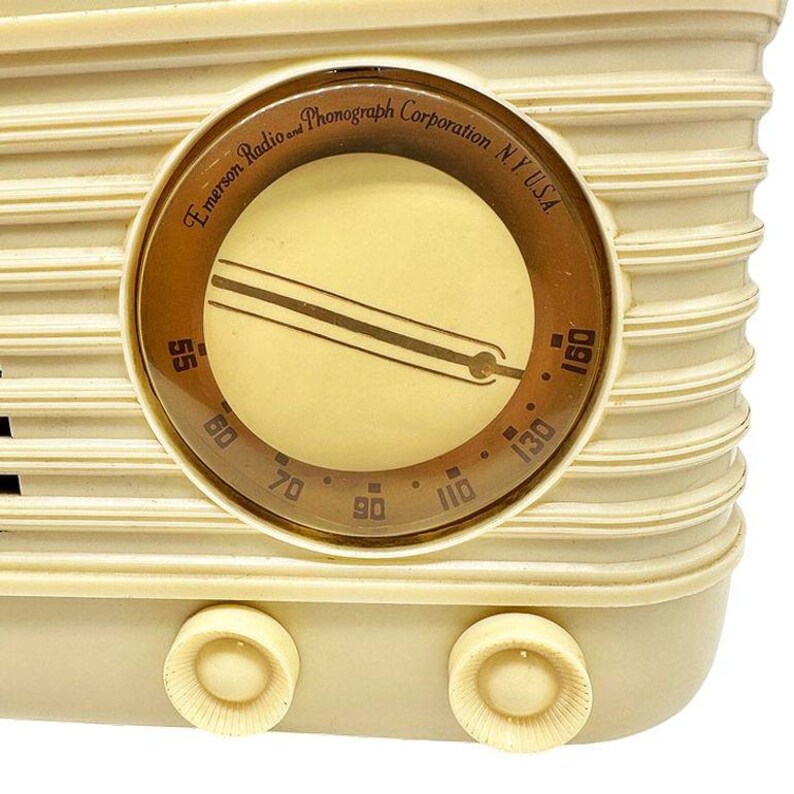 Puede incluir: Primer plano de una radio vintage de color crema con un dial circular. El dial presenta un anillo exterior marr&oacute;n con el texto "Emerson Radio and Phonograph Corporation N.Y. USA". El dial muestra n&uacute;meros del 55 al 160. Se ven dos perillas de color crema.