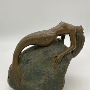 May include: A bronze sculpture of a mermaid reclining on a rock. The mermaid has a long, flowing tail and is looking up towards the sky. The sculpture is detailed and realistic.