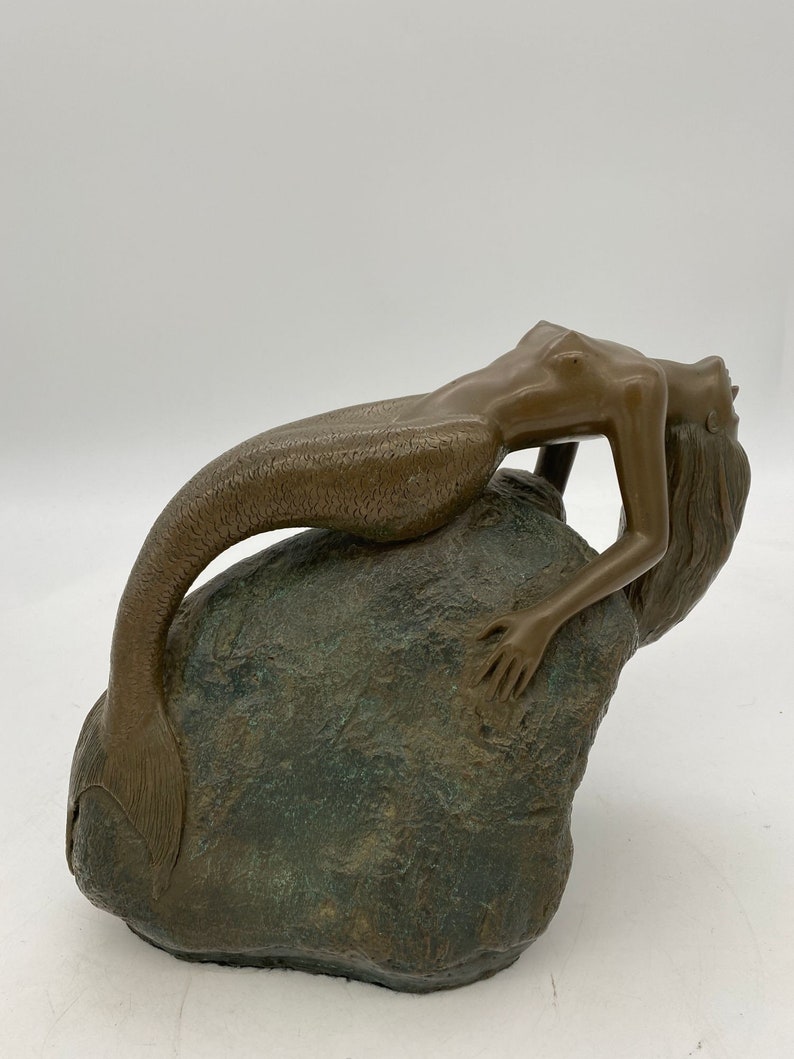 May include: A bronze sculpture of a mermaid reclining on a rock. The mermaid has a long, flowing tail and is looking up towards the sky. The sculpture is detailed and realistic.