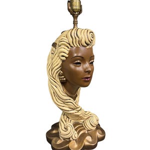 May include: A ceramic table lamp shaped like a woman's head with blonde hair and a gold base.