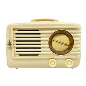 Puede incluir: Radio vintage de color crema con rejilla de altavoz, dial y mandos. La radio tiene un dise&ntilde;o retro con un asa en la parte superior y un dial circular con n&uacute;meros. El texto "Detrola" es visible.