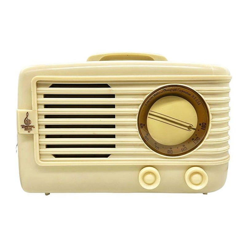 Puede incluir: Radio vintage de color crema con rejilla de altavoz, dial y mandos. La radio tiene un dise&ntilde;o retro con un asa en la parte superior y un dial circular con n&uacute;meros. El texto "Detrola" es visible.