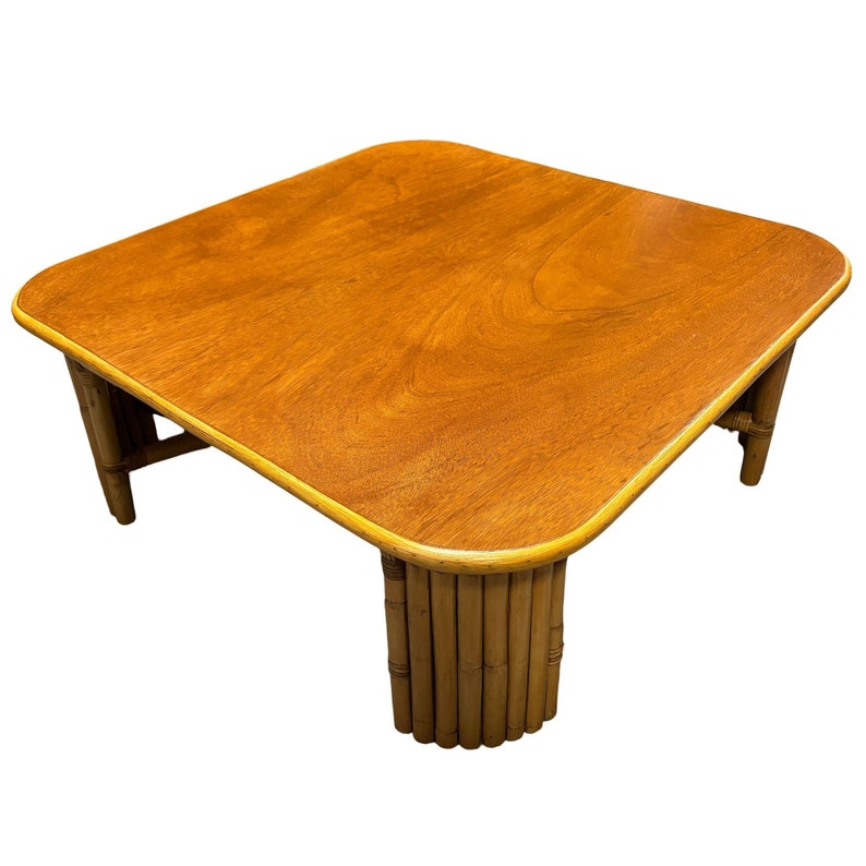 Pode incluir: Uma mesa de centro quadrada com tampo de madeira marrom claro e base de bambu. A mesa tem uma borda arredondada e uma ligeira borda elevada ao redor do tampo.