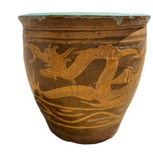 May include: A brown ceramic pot with a light green rim and a detailed dragon design in a lighter brown color. The pot has a wide, rounded shape and a textured surface, showcasing an antique aesthetic.