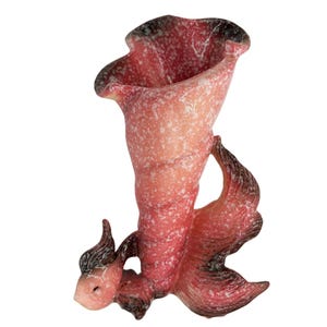 May include: A decorative art glass vase in the shape of a fish. The vase is primarily pink with white speckles and dark brown accents. The flared top and tail fins add to the unique design. A whimsical piece for home decor.