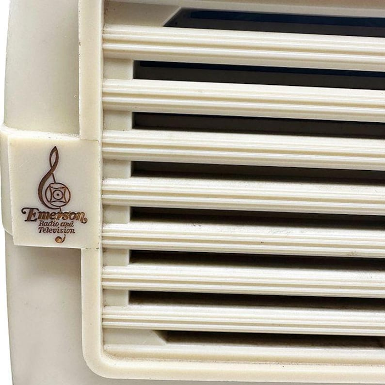Puede incluir: Primer plano de una radio Emerson vintage. La radio tiene una carcasa de pl&aacute;stico color crema con ranuras de ventilaci&oacute;n horizontales. El logotipo de Emerson, con el nombre de la marca y un dise&ntilde;o estilizado, es visible en el lateral.