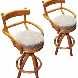 May include: Two brown bamboo bar stools with a round, light gray, patterned cushion on each seat. The stools have a curved back and a circular footrest.
