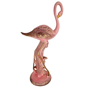 May include: Pink ceramic flamingo figurine with gold accents. The flamingo has a long, curved neck and is perched on a decorative base. The figurine has a textured, glittery finish, adding a touch of sparkle. A decorative piece for a shelf or table.