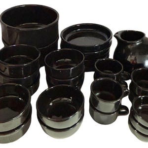 May include: A collection of glossy black ceramic dinnerware including large bowls, small bowls, saucers, a pitcher with a handle, and several mugs. The set features a uniform dark finish across all pieces, which are arranged in stacks and groups.