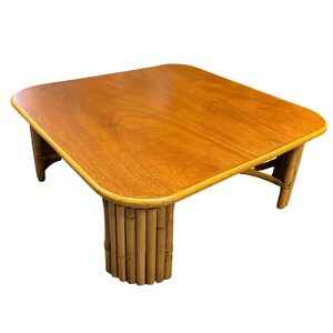 Pode incluir: Uma mesa de centro quadrada com um tampo de madeira marrom claro e uma borda arredondada. A mesa tem uma base &uacute;nica feita de bambu tecido.