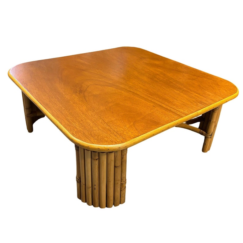 Pode incluir: Uma mesa de centro quadrada com um tampo de madeira marrom claro e uma borda arredondada. A mesa tem uma base &uacute;nica feita de bambu tecido.