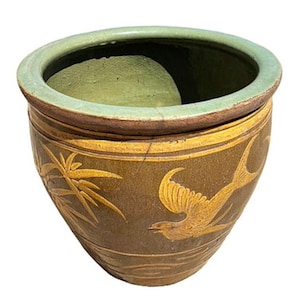 May include: A round, glazed ceramic planter with a wide opening and a slightly tapered body. The planter is primarily brown with a green rim and features gold-colored etched designs of a bird and bamboo.