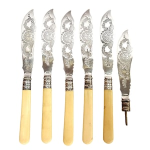May include: Set of six antique butter knives. The blades are silver-colored with an engraved floral design. The handles are a creamy ivory color, and the knives have a silver-colored collar. One knife has a different handle and a small, pointed end.