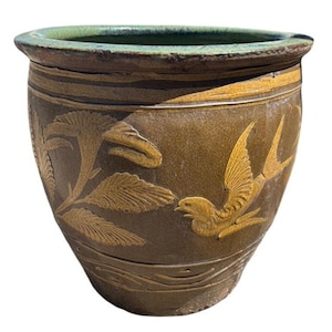 May include: A round, brown ceramic planter with a green rim. The planter features embossed gold-colored designs of a bird and foliage. The pot has a wide opening and a slightly textured surface, suitable for indoor or outdoor use.