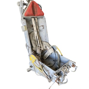 May include: A vintage aircraft ejection seat with a gray metal frame, red padded headrest, and yellow and white straps. The seat has a worn and weathered appearance.