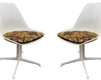 May include: Two white, modern chairs with a curved back and a four-legged base. The seat cushions feature a colorful pattern of green leaves and purple grapes, adding a touch of nature-inspired design to the furniture.