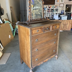 May include: A vintage wooden dresser with a beveled mirror on top. The dresser has five drawers with ornate brass hardware and a carved design on the bottom. The dresser is a rich brown color and has turned legs.