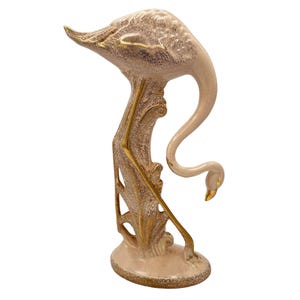 May include: A ceramic flamingo sculpture in a cream and gold color scheme. The flamingo is depicted with its head gracefully curved downward, with gold accents on the beak, legs, and decorative base. The sculpture has a textured finish.