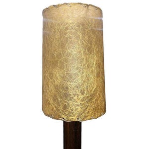 May include: A vintage lampshade with a beige, textured fabric. The shade has a woven, fringed edge and a brown wooden base.