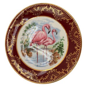 May include: A decorative plate featuring a central image of two pink flamingos standing in water, surrounded by greenery. The plate has a burgundy border with gold filigree and a gold rim, creating an elegant aesthetic.