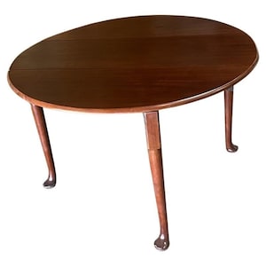 May include: A round, wooden, drop-leaf table with a dark brown finish. The table has four legs with cabriole legs and a carved foot.