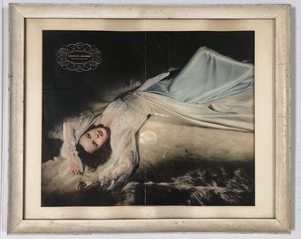 May include: A framed vintage advertisement featuring a woman in a light blue satin gown, lying on a fur-like surface. The image has a soft, romantic feel, with a logo at the top left corner. The frame is a distressed, off-white color.