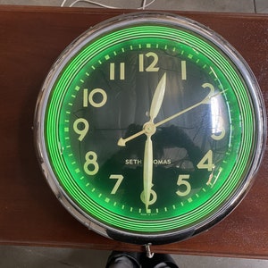 May include: A vintage green neon clock with a chrome frame. The clock face has black numbers and a black background. The clock says "Seth Thomas" on the face.