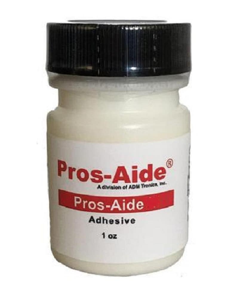 Op de afbeelding: Een kleine, witte fles Pros-Aide lijm met een zwarte dop. Het etiket toont de merknaam in rood en wit, samen met de tekst "Adhesive" en "1 oz" (ongeveer 28 g).
