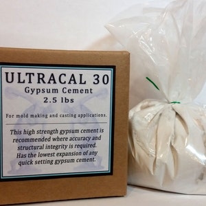 Op de afbeelding: Een bruine kartonnen doos en een doorzichtige plastic zak gevuld met witte gipsmortel. De doos heeft een label met de tekst "ULTRACAL 30 Gypsum Cement 1.13 kg" en is voor het maken van mallen en gieten.