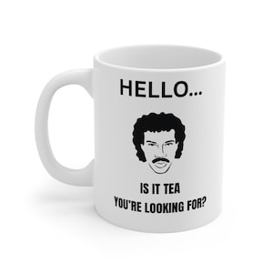 以下が含まれることがあります： 白いセラミックマグカップ。黒い男性の顔のグラフィックと「HELLO... IS IT TEA YOU'RE LOOKING FOR？」のテキストがプリントされています。マグカップにはカーブしたハンドルがあり、温かい飲み物用に設計されています。
