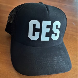 May include: A black trucker hat with a white mesh back and the word "CES" in white block letters on the front.