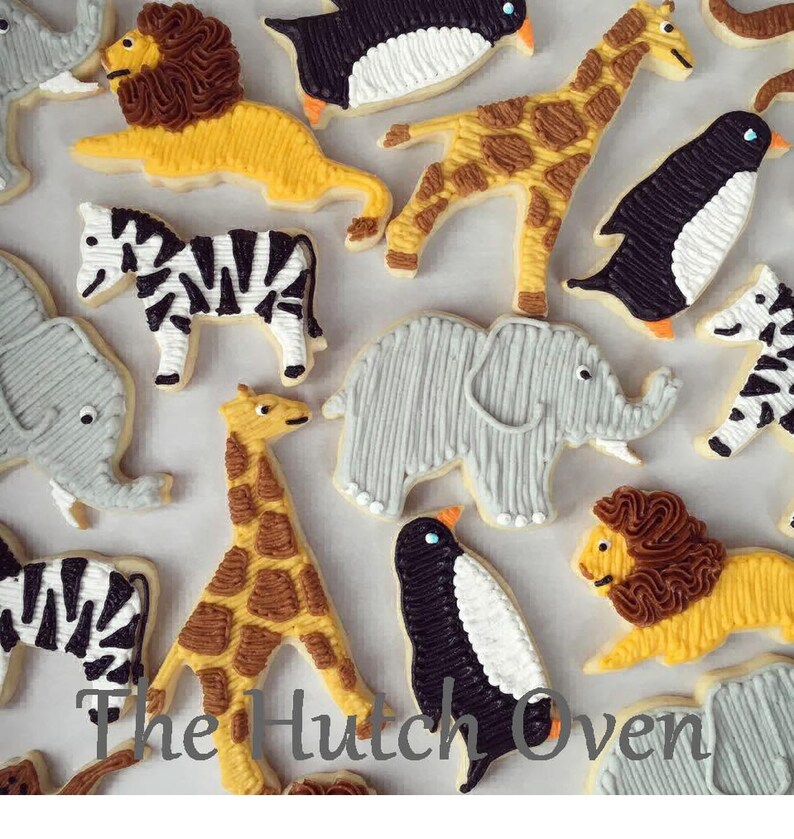 May include: A collection of decorated sugar cookies featuring various animal designs, including a zebra, giraffe, penguin, lion, and elephant. The cookies are decorated with intricate details and bright colors.