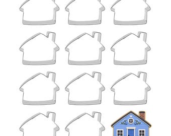 May include: Eleven silver-coloured metal house-shaped cookie cutters arranged in a grid, accompanied by one finished house-shaped cookie decorated with blue icing, a blue door, white windows, a chimney, and small floral accents around the exterior.
