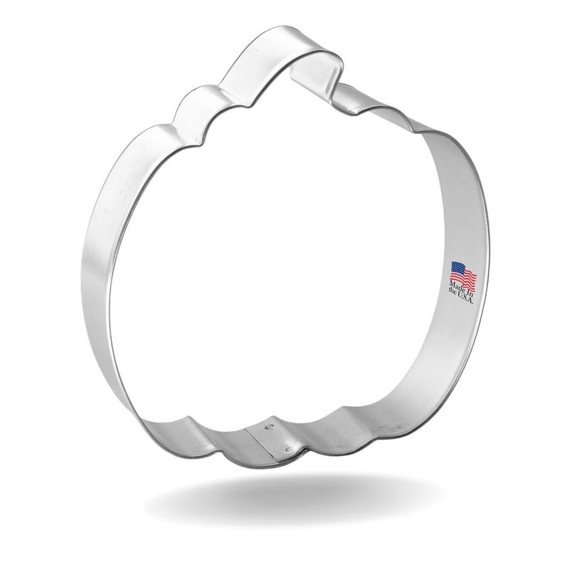 May include: A shiny, silver-colored pumpkin-shaped cookie cutter. The cutter has a scalloped edge and a curved handle at the top. The words "Made in the USA" are printed on the side with a small American flag.