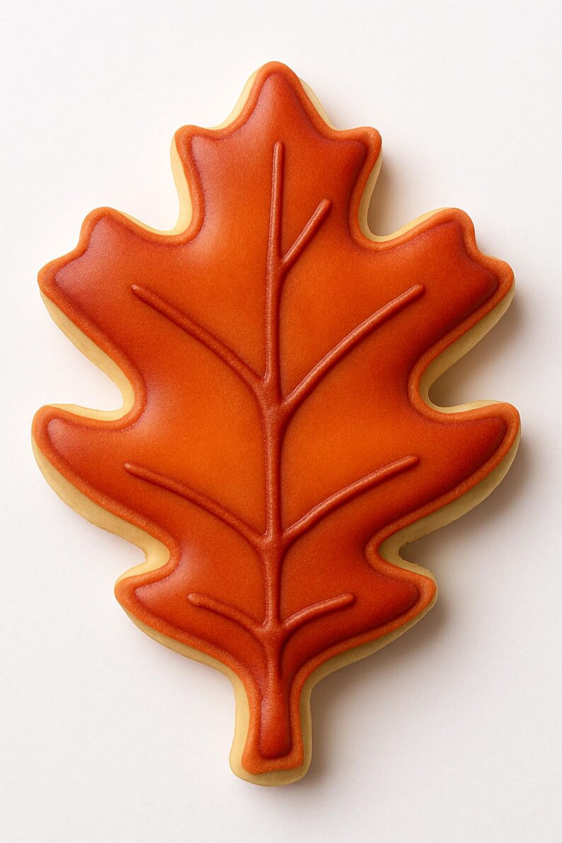 May include: A leaf-shaped cookie with orange icing and a detailed vein design. The cookie has a light-colored edge, and the orange icing is smooth and glossy. This autumn-themed treat is perfect for fall celebrations.