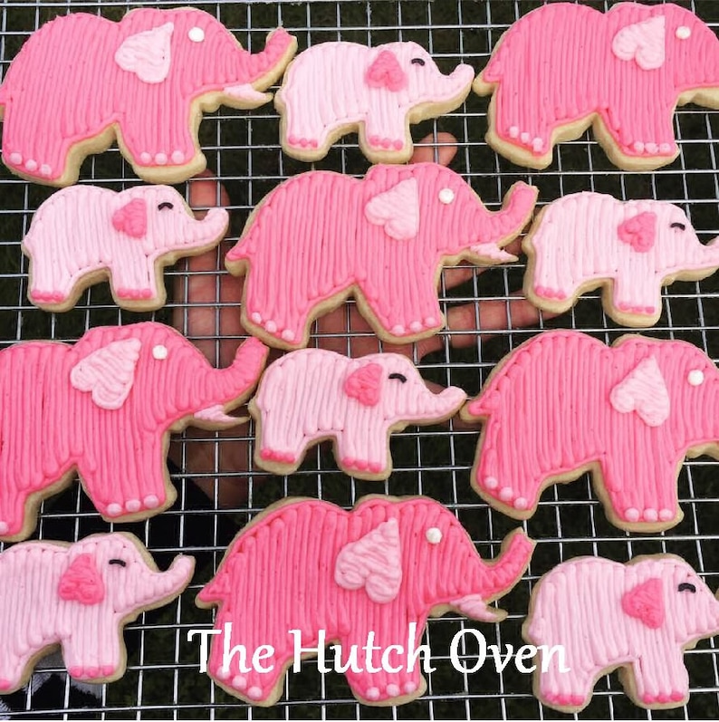 May include: A collection of pink elephant-shaped sugar cookies decorated with pink frosting and white dots. The cookies are arranged on a wire rack. The text "The Hutch Oven" is visible in the bottom center of the image.