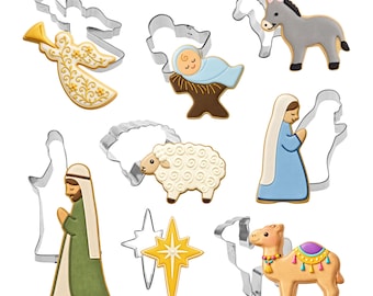 May include: A collection of eight metal cookie cutters paired with matching decorated cookies featuring a nativity scene. The set includes an angel, a baby in a manger, a donkey, a sheep, a figure in a blue robe, a figure in a green robe, a star, and a camel.