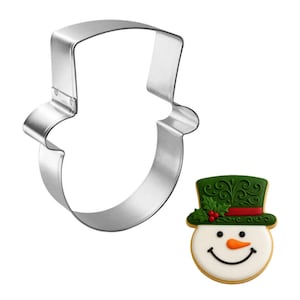 May include: A stainless steel snowman-shaped cookie cutter next to a decorated snowman cookie. The cookie has a white face, a green hat with red accents, and a smiling face with a carrot nose. Perfect for holiday baking.