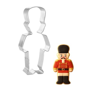 May include: A cookie cutter and a decorated cookie in the shape of a toy soldier. The cookie cutter is made of silver metal. The cookie is decorated with red, black, and tan icing, and has a black hat and mustache.