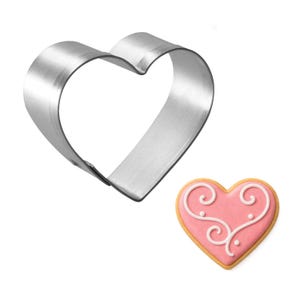 May include: A shiny, silver heart-shaped cookie cutter next to a decorated heart-shaped cookie. The cookie has a pink frosting base with white icing swirls and a yellow-brown border. The cutter is made of metal.