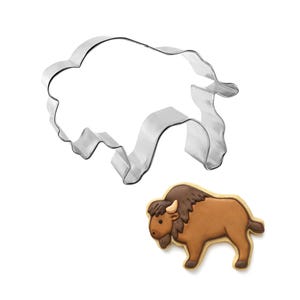 May include: A silver-colored metal cookie cutter shaped like a bison, positioned above a single sugar cookie decorated with brown and tan icing to resemble a bison with a dark brown mane, a small horn, and a short tail, all on a white background.