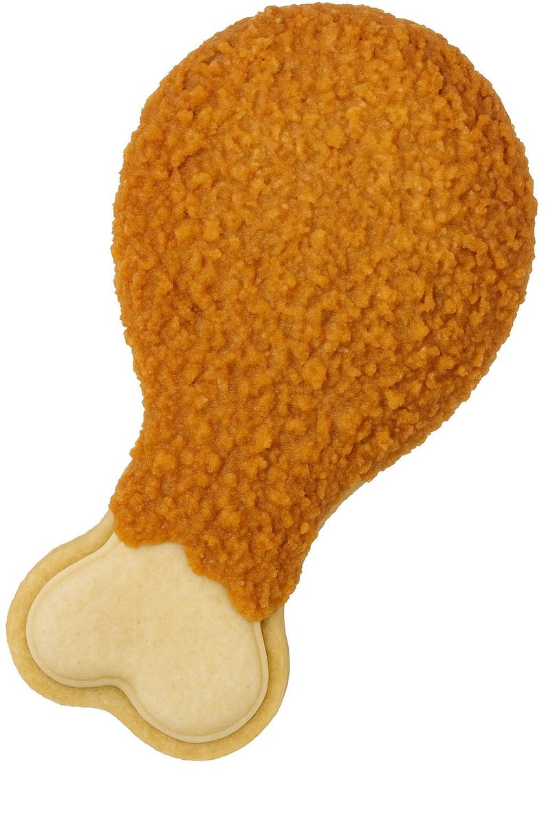 May include: A cookie shaped like a fried chicken leg. The top portion is covered in a textured, golden-brown coating, while the bottom is a light tan color, resembling the bone. The cookie is set against a plain white background.