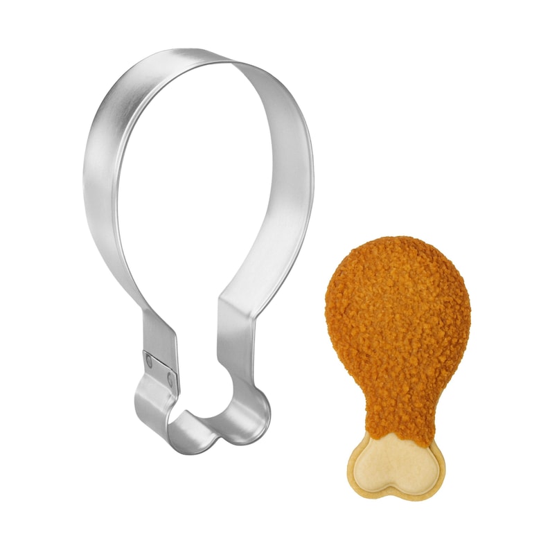 May include: A stainless steel cookie cutter shaped like a fried chicken leg next to a baked cookie in the shape of a fried chicken leg. The cookie has a golden-brown, textured coating and a light-colored bone.